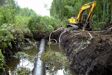 Big yellow excavator near a pit with a gas pipeline. The pit is filled with water. Forest in the...