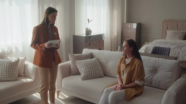 Female buyer sitting on white sofa clarifying things about pillows and couch with furniture store manager
