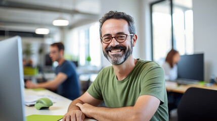 Cheerful man with glasses and a green t-shirt is smiling at the camera, seated in a modern office with co-workers and computers in the background. - obrazy, fototapety, plakaty