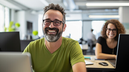 Cheerful man with glasses and a green t-shirt is smiling at the camera, seated in a modern office with co-workers and computers in the background.