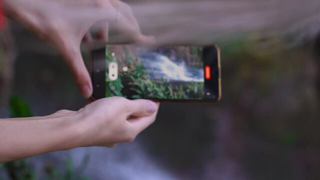 tourist taking pictures amazing waterfall on smartphone Close-up of female tourist's hands shooting video of waterfall on smartphone on green natural landscape background. Tourist vacation