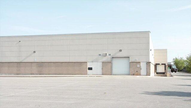 white doors and receiving bay doors on large one story beige building with large empty parking cement lot pavement space in front, middle of frame, medium wide shot