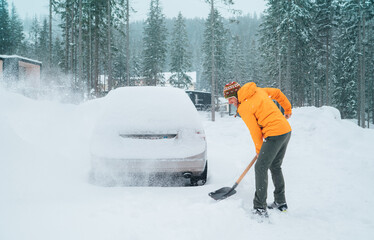 Smiling man with a shovel removing snow from the path and cleaning auto. Car covered with snow as a...