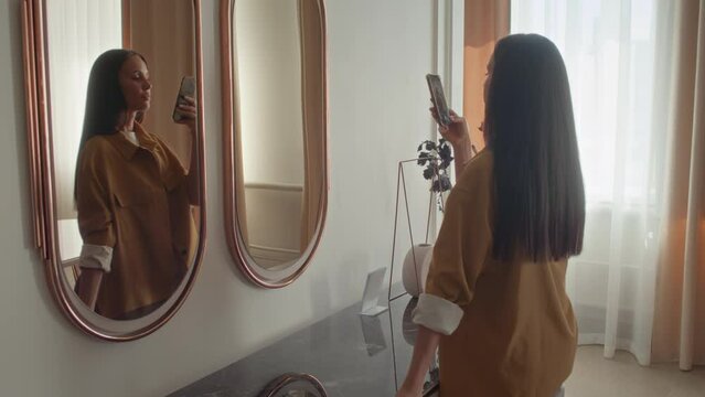 Caucasian woman with dark hair coming to mirror, taking couple of photos in her reflection and walking away