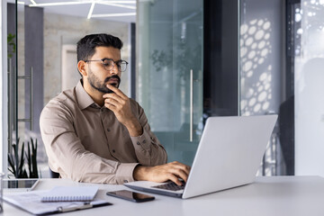 Man thinking inside office at workplace with laptop, businessman preparing financial report, typing on keyboard, financier accountant in glasses and shirt holding hand to chin.