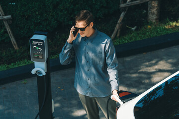 Young man travel with EV electric car charging in green sustainable city outdoor garden in summer...