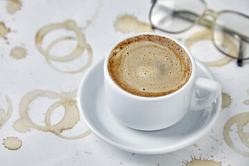 White cup of coffee on a saucer and reading glasses on a table stained with imprints of coffee cups