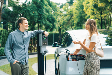 Young couple travel with EV electric car charging in green sustainable city outdoor garden in...