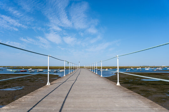 Empty walkway over blue ocean, coastline and horizon seen a large long wooden jetty with a thin white metal railing along the edges but in the perspective distance a blue sky and an ocean with boats.