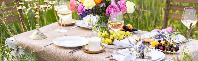 Beautiful romantic outdoor wedding decor in a field. Table decorated with burning candles, flowers. Wineglasses with white wine. Sunset, summer, golden hour. Perfect surprise date for couple. Banner