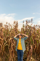 granjera con gran sonriza con la mano sujetando su sombrero de paja lista para lanzarlo por los aires esta parada frente a un sembrío de cañas de maíz seco, mujer adulta en sus 50 años feliz y empoder - obrazy, fototapety, plakaty