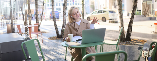 Portrait of young blond woman, female student in street cafe, wearing wireless headphones, using laptop, having online meeting, attend web lecture or course, doing homework - Powered by Adobe