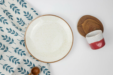 Composition of kitchen utilities: white plate, purple and white cup, wooden items on textile leaves pattern background, top view