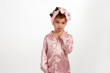 Cute little girl with curlers and a pink headband, wearing pink silk pajamas on a white background.