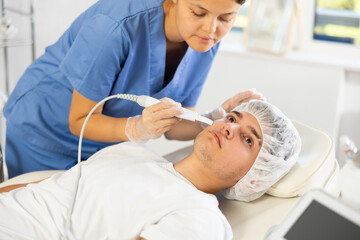 Young female cosmetologist performs hardware facial exfoliation procedure to young male patient