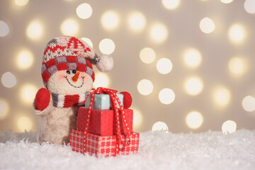 Toy snowman with gift box and decorative Christmas tree on snow and glowing lights bokeh...