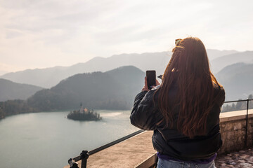 Woman tourist at beautiful Lake Bled with Church on an island taking a photograph with a smartphone
