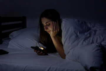 Fototapeta na wymiar A girl in bed with a phone on a white bed in a dark room.