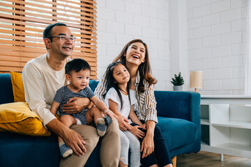 In the heart of their modern grooved house a joyful Asian family enjoys quality family time watching TV together on the cozy sofa. Laughter smiles and togetherness define their weekend.