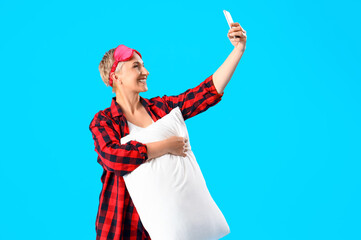 Fototapeta na wymiar Mature woman in pajamas with pillow taking selfie on blue background