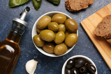 Bowls with ripe olives and bottle of oil on blue background