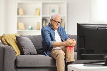 Mature man in pain sitting on a sofa in front of tv and holding his inflamed knee - obrazy, fototapety, plakaty