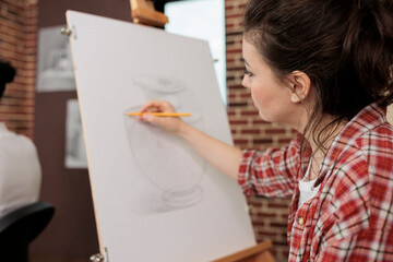 Young woman sitting at easel sketching basic vase on canvas using pencil, student girl learning...