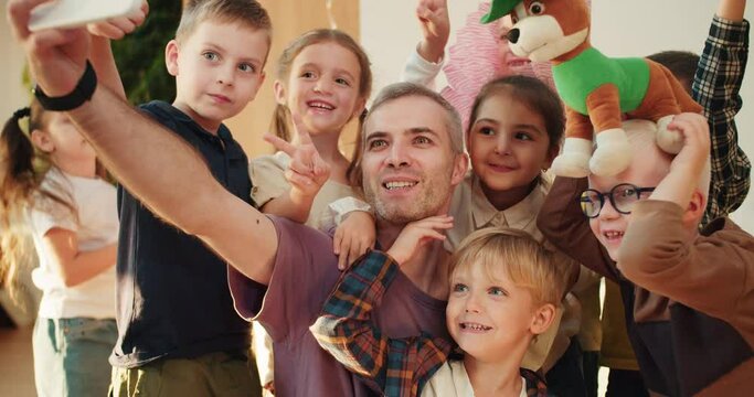 A group of happy preschool children together with their teacher, a man with gray hair in a purple T-shirt, take a selfie at their lesson in a club for preparing children for school