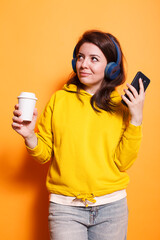 Portrait of a relaxed woman in a studio holding a recyclable coffee cup and listening to music on wireless headphones. Female individual holding smartphone is using an online streaming app.