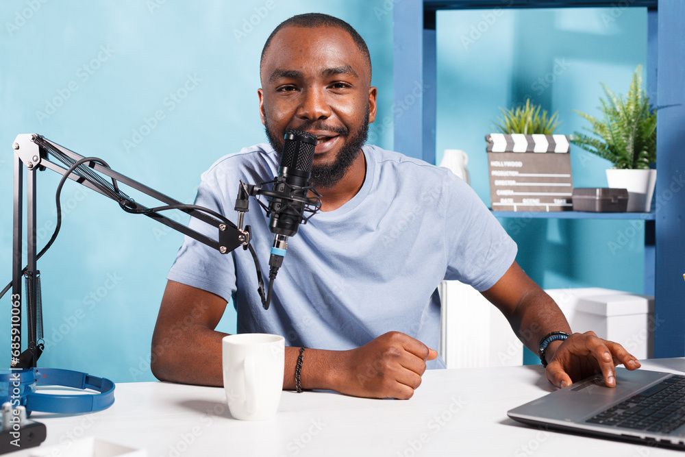 Poster smiling vlogger speaking in microphone, using laptop and looking at camera while recording video for