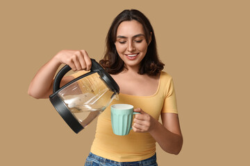 Beautiful young woman pouring hot water from modern electric kettle into cup on beige background