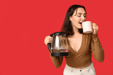 Beautiful young woman with modern electric kettle drinking coffee on red background
