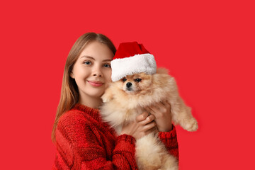 Fototapeta premium Young woman with cute Pomeranian dog in Santa hat on red background