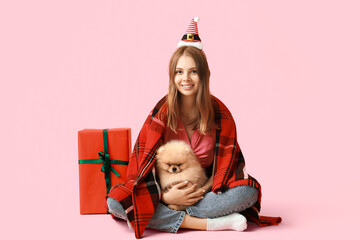 Young woman with cute Pomeranian dog in reindeer horns and Christmas gift box on pink background