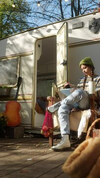An attractive hippie girl sketching on a tablet while seated in front of the trailer in the autumn garden.