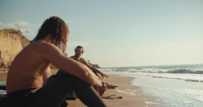 Three surfer guys with naked torsos sit on the seashore on sandy beach and communicate with each other in wetsuits on the beach in summer