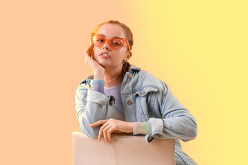 Beautiful young woman with stylish sunglasses sitting on chair against color background