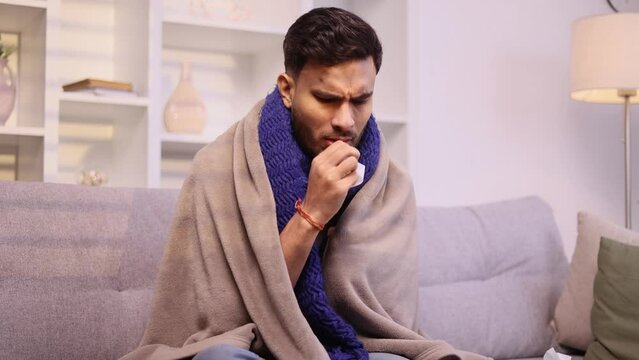 Sick unhealthy young man on couch with warm scarf coughing sneezing and having severe headache temperature indoors Sad tired guy getting flu virus symptom at cold season at home