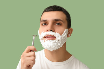 Handsome young man with razor and shaving foam on his face against green background