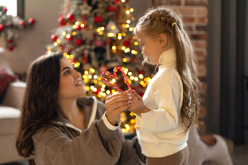 Little girl and her mom sitting near Christmas tree, have fun and hugging. Hands of mother and child girl holding heart-shaped candy on festive Christmas tree background