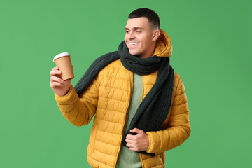 Young man in yellow stylish puffer jacket and scarf with paper cup of coffee on green background
