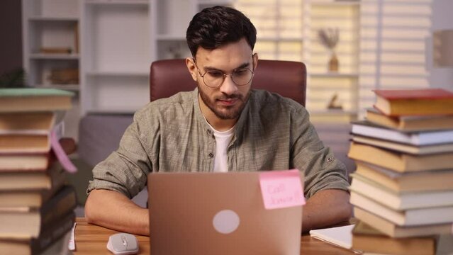 Focused motivated young handsome man student with stack of books typing browsing studying on laptop computer and looking at camera at home office Distance remote education concept