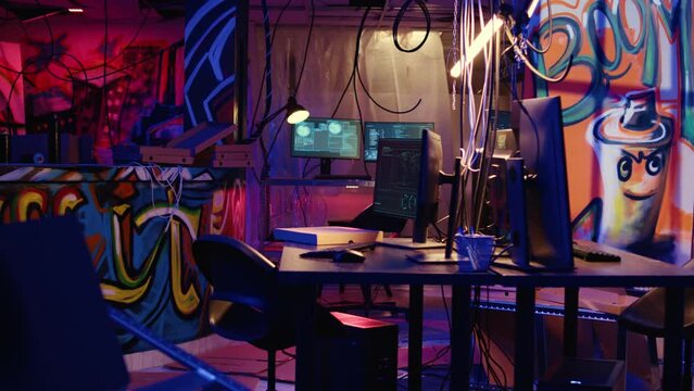 Dolly out shot of empty messy hackers base of operations with neon lights and graffiti drawings sprayed on walls. Ghetto hideout used by criminals to commit illegal activities using computers