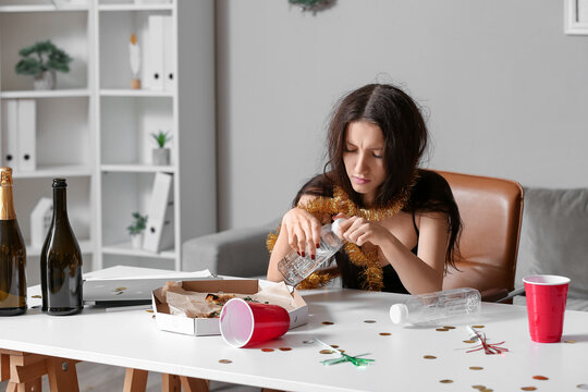 Young woman with hungover drinking water at table in office after New Year party