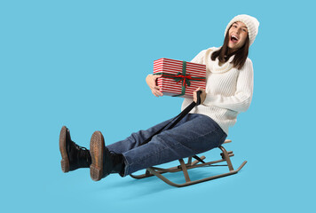 Young woman with Christmas gift sledging on blue background