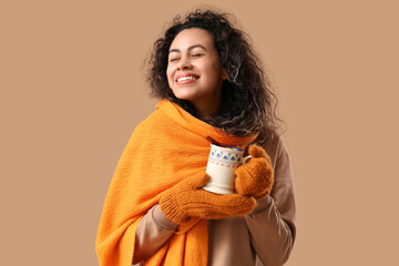 Happy young African-American woman in warm mittens with cup of hot mulled wine on brown background