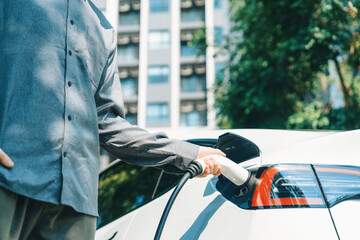 Young man travel with EV electric car charging in green sustainable city outdoor garden in summer...