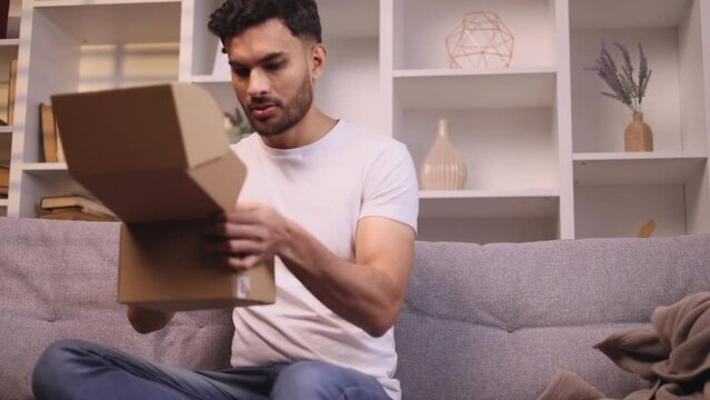 Portrait of young handsome man received parcel dissatisfied with shopping in online internet store at home Upset guy customer disappointed with purchase order while open carton box indoors