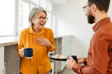 Bearded man giving cookies plate to senior woman colleague while enjoying coffee break