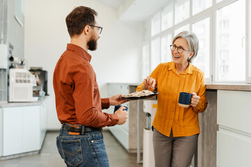 Friendly man giving cookies plate to senior woman colleague while enjoying coffee break
